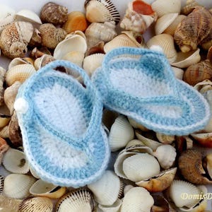 May include: A pair of blue and white crocheted baby sandals, resembling flip-flops, are displayed on a bed of seashells.