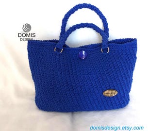 May include: A royal blue crocheted tote bag with a gold button and a gold oval tag that says "DOMIS DESIGN".