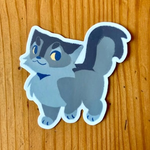 Puede incluir: Una pegatina con un gato de dibujos animados con una cola esponjosa. El gato es principalmente gris y azul con ojos amarillos y un peque&ntilde;o lazo azul. La pegatina est&aacute; sobre una superficie de madera.