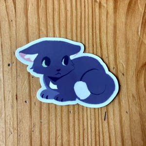 Puede incluir: Una pegatina con un gato de dibujos animados en tonos azules. El gato tiene ojos grandes, orejas rosas peque&ntilde;as y una mancha blanca en la parte trasera. La pegatina est&aacute; sobre un fondo de madera.