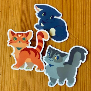 Puede incluir: Tres pegatinas de gatos con ilustraciones de dibujos animados. Una es naranja con una cola a rayas, otra es azul oscuro y la tercera es gris. Cada pegatina tiene un borde blanco y est&aacute; colocada sobre una superficie de madera.
