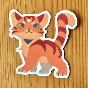 Puede incluir: Una pegatina con un gato naranja estilizado con rayas rojas oscuras y ojos verdes. El gato tiene un contorno blanco y un peque&ntilde;o collar azul. La pegatina est&aacute; sobre una superficie de madera.