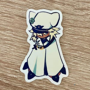 May include: A die-cut sticker featuring a cartoon character in a white outfit with a matching hat. The character has blonde hair, a black eye mask, and a gold belt. The sticker has a white border and is on a wooden surface.