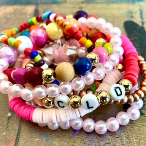 May include: A stack of colorful beaded bracelets with pink, red, blue, yellow, and white beads. The bracelets spell out the word "COLD" in white letters on black beads.