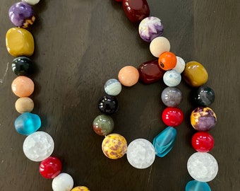 Op de afbeelding: Een kleurrijke kralenketting met verschillende stenen en glazen kralen. De ketting heeft een mix van kleuren, waaronder rood, oranje, geel, groen, blauw, paars en wit.