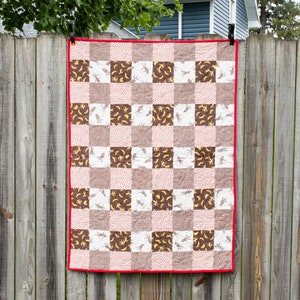 May include: A brown and white checkered baby quilt with a red border. The quilt features a repeating pattern of small brown monkeys and yellow bananas.