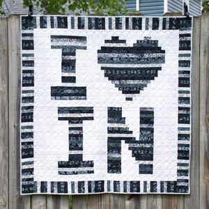 May include: A handmade quilt with the words "I" and "IN" and a heart shape in the middle. The letters and heart are made of black and gray patterned fabric on a white background. The quilt is hanging on a wooden fence.