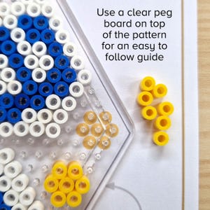May include: A close-up of a clear pegboard with a blue and white bead pattern. Yellow beads are arranged in a flower shape on the board. The text "Use a clear peg board on top of the pattern for an easy to follow guide" is visible in the background.