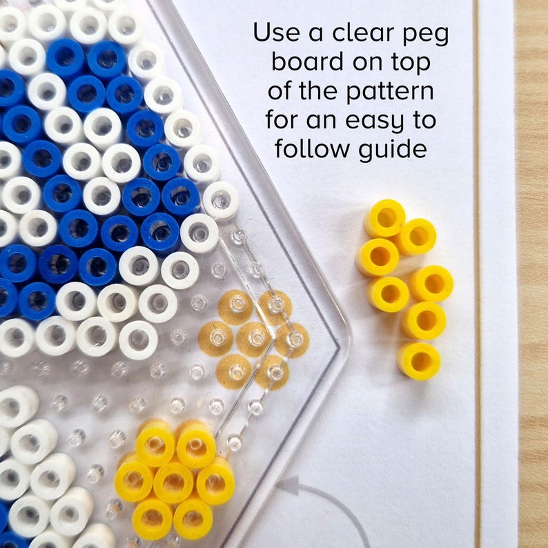 May include: A close-up of a clear pegboard with a blue and white bead pattern. Yellow beads are arranged in a flower shape on the board. The text "Use a clear peg board on top of the pattern for an easy to follow guide" is visible in the background.