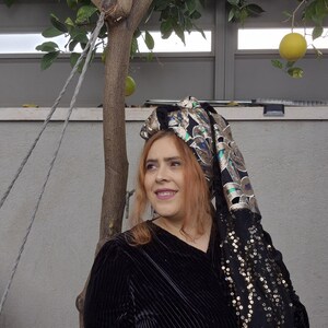 Puede incluir: Una mujer con un vestido de terciopelo negro y un pañuelo de cabeza con lentejuelas doradas y verdes. El pañuelo tiene un estampado floral y está anudado en la parte posterior de la cabeza.