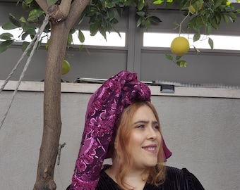 May include: A woman wearing a black velvet dress with a purple sequined head scarf. The scarf is tied in a knot on top of her head and has a floral pattern. She is standing in front of a tree with green leaves and yellow fruit.