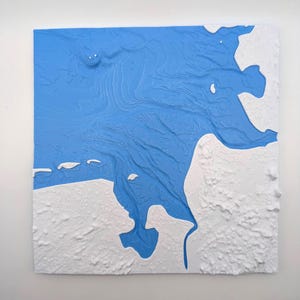 Puede incluir: Una obra de arte cuadrada y texturizada con un diseño topográfico. La pieza presenta una zona central azul brillante, que se asemeja al agua, rodeada por un borde blanco texturizado. El diseño en relieve añade profundidad e interés visual a la obra.