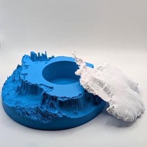 Pode incluir: Modelo impresso em 3D da Antártida, com uma base azul e uma peça branca separada que representa o continente. A peça branca mostra linhas topográficas detalhadas. O modelo está sobre uma superfície branca.
