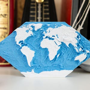 Op de afbeelding: Een diamantvormig decoratief object met een wereldkaart. De continenten zijn wit en de oceanen blauw. Dit is een uniek woondecoratie-item met een getextureerd oppervlak.
