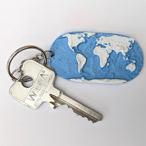 May include: A silver key with the text "WILKA SCHLIESSTECHNIK" attached to a keychain next to a blue and white world map keychain. The world map keychain is an oval shape with a textured blue background and white continents.
