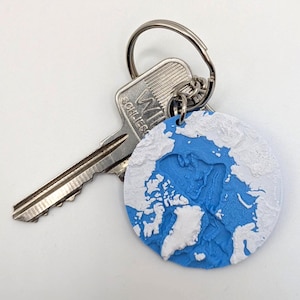 Peut inclure: Un porte-clés rond représentant une carte en relief 3D de l'Arctique, avec des océans bleus et des terres blanches. Le porte-clés est attaché à une clé argentée et à un anneau.