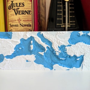 Może przedstawiać: Prostokątna mapa reliefowa Morza Śródziemnego, z białymi lądami i niebieskim morzem. Mapa jest wyświetlana na białej powierzchni. W tle widoczne są książki i metronom. Mapa jest prawdopodobnie wykonana z gipsu.