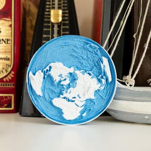 May include: A circular decorative item featuring a world map design in blue and white. The continents are white against a textured blue backdrop. It's displayed with books and a small model sailboat.