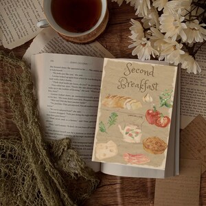 Puede incluir: Un libro abierto con una ilustración de acuarela de una escena de desayuno. La ilustración presenta pan, queso, tocino, tomates, ajo y un pastel. El título del libro es "Second Breakfast".