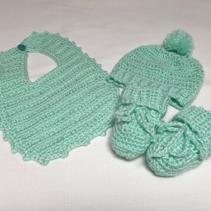 May include: A set of crocheted baby accessories in a light green color. The set includes a bib, a hat with a pom-pom, and booties.