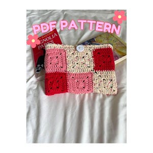 May include: A handmade crochet book sleeve featuring a patchwork design in red, pink, and cream squares. The sleeve is secured with a white button. The text "PDF PATTERN" is displayed in pink letters above the sleeve.