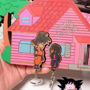 May include: A set of three wooden keychains featuring characters from the anime Dragon Ball Z. The keychains are painted in bright colors and depict Goku, Bulma, and the Kame House. The keychains are attached to silver key rings.