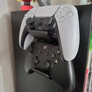 Peut inclure: Deux manettes de jeu vidéo montées sur un mur. La manette du haut est blanche avec des boutons noirs et la manette du bas est noire avec des boutons verts, jaunes et rouges.