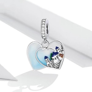 May include: Silver charm in the shape of two interlocking hearts. The charm features a white enamel background with a blue and white painted scene of a small town with white buildings and blue roofs. The charm has a silver loop at the top for attaching to a bracelet.