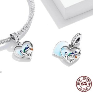 May include: A silver charm in the shape of two interlocking hearts. The hearts are open and feature a white enamel background with a colourful illustration of a building. The charm is attached to a silver chain.