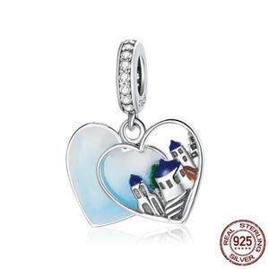 May include: Silver charm in the shape of two interlocking hearts. One heart features a white and blue enamel design of a small town with buildings. The other heart is a solid blue enamel.