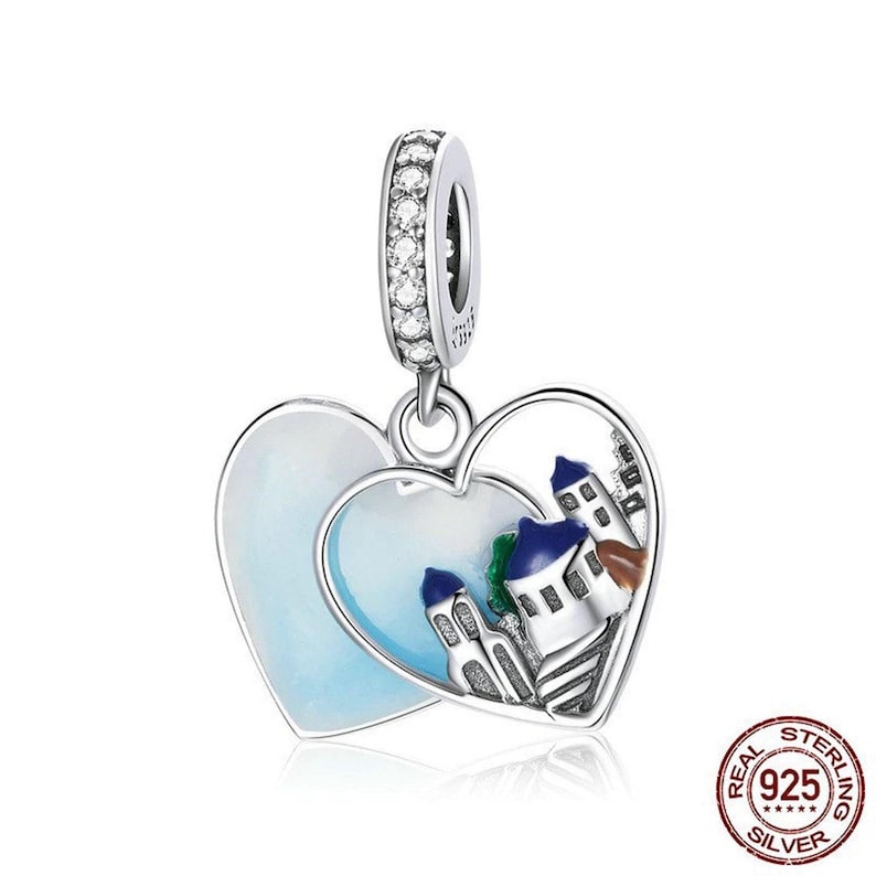 May include: Silver charm in the shape of two interlocking hearts. One heart features a white and blue enamel design of a small town with buildings. The other heart is a solid blue enamel.