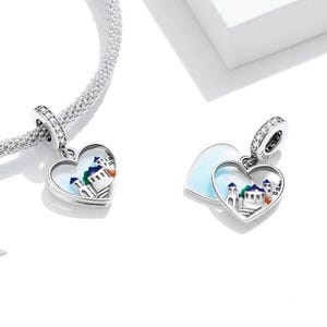 May include: Two silver heart-shaped charms with a white enamel background and a colourful painted scene of a building with a blue roof and white walls. The charms are attached to a silver chain.