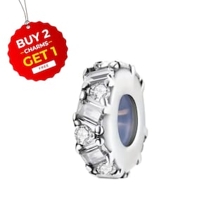 May include: A silver charm featuring clear gemstones and a central blue-tinted hole. The charm is decorated with round and rectangular-cut stones. A red tag with the text "BUY 2 CHARMS GET 1 FREE" is visible.