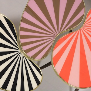 May include: Three connected, abstract, kidney-shaped tables with gold trim. The tabletops are painted with a sunburst pattern in black and white, pink and brown, and pink and orange.