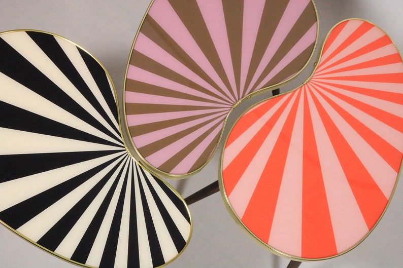 May include: Three connected, abstract, kidney-shaped tables with gold trim. The tabletops are painted with a sunburst pattern in black and white, pink and brown, and pink and orange.