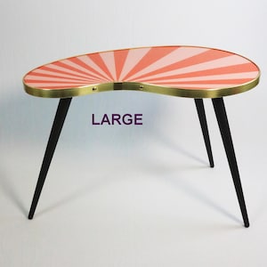Può includere: Tavolino a forma di rene in stile retrò. Il piano presenta un motivo a raggi di sole corallo e bianco, bordato da un bordo metallico dorato. Ha tre gambe nere affusolate. La parola "LARGE" è stampata sul tavolo.