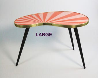 May include: A mid-century modern kidney-shaped table. The tabletop features a coral and white sunburst design with a gold-colored metal trim. The table has three black, tapered legs. The word "LARGE" is printed on the table.