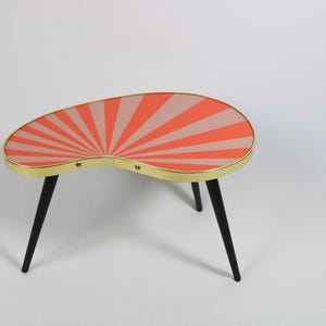 May include: A kidney-shaped coffee table featuring a vibrant sunburst pattern in alternating shades of coral and light pink. The tabletop is encased in a thin gold-toned metal rim and is supported by three slender, tapered black legs.