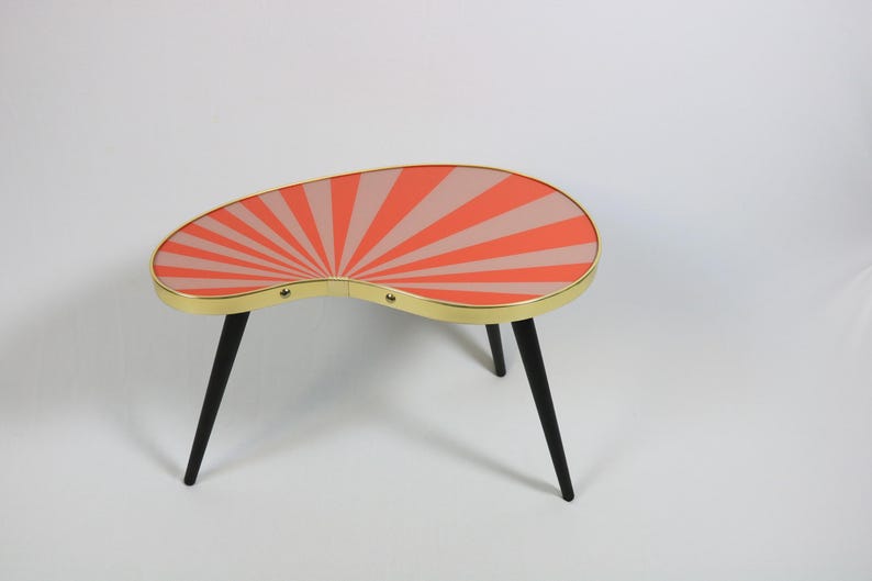 May include: A kidney-shaped coffee table featuring a vibrant sunburst pattern in alternating shades of coral and light pink. The tabletop is encased in a thin gold-toned metal rim and is supported by three slender, tapered black legs.