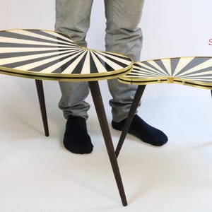 May include: Two mid-century modern side tables. The tables have a black and white sunburst design on the oval tabletop, with a gold trim. The tables have dark brown, tapered legs. The larger table is approximately 30 inches long.