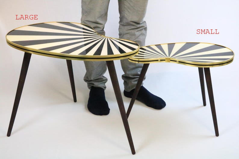May include: Two mid-century modern side tables. The tables have a black and white sunburst design on the oval tabletop, with a gold trim. The tables have dark brown, tapered legs. The larger table is approximately 30 inches long.