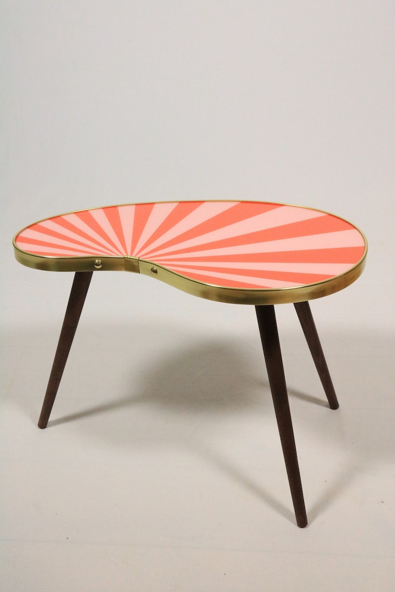 May include: A kidney-shaped coffee table featuring a tabletop with a radiating sunburst pattern in alternating shades of coral and light pink. The tabletop is edged with a metallic gold band and supported by three tapered dark brown wooden legs.