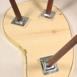May include: The underside of a wooden table with a unique, asymmetrical shape. The table has a brass trim around the edge and three wooden legs with metal brackets.