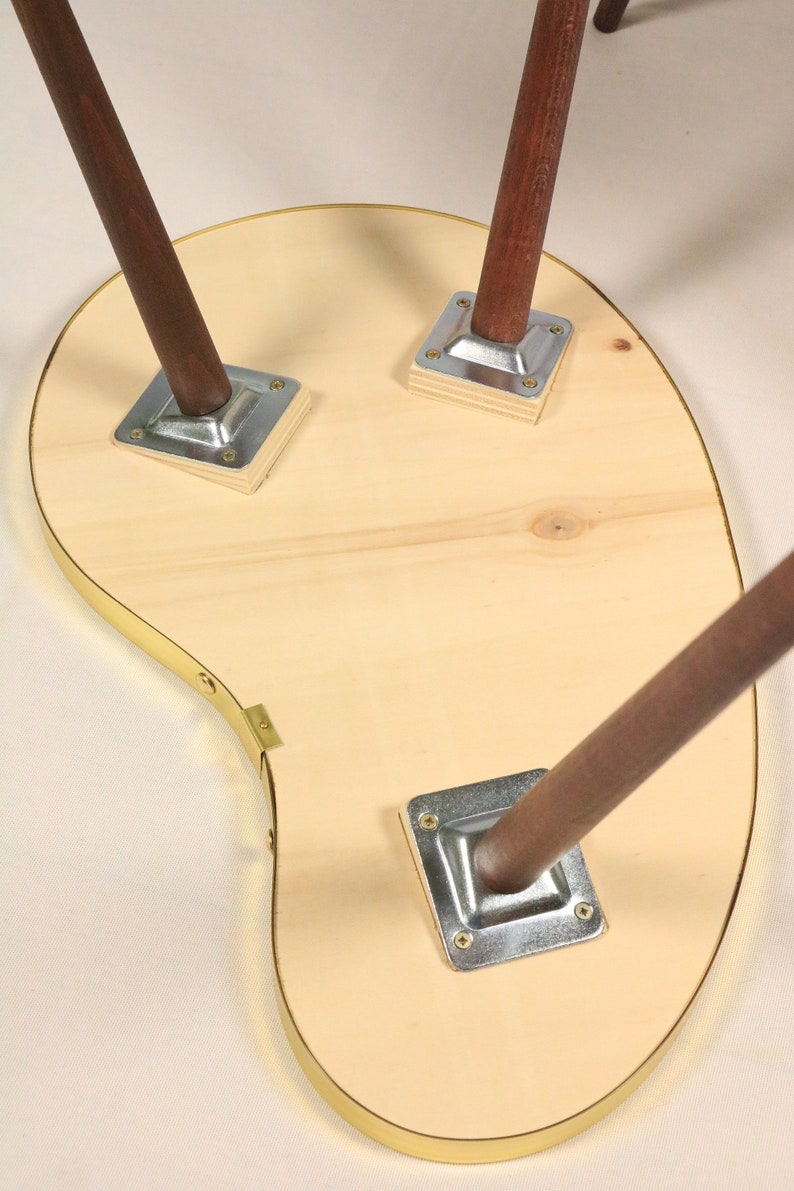 May include: The underside of a wooden table with a unique, asymmetrical shape. The table has a brass trim around the edge and three wooden legs with metal brackets.