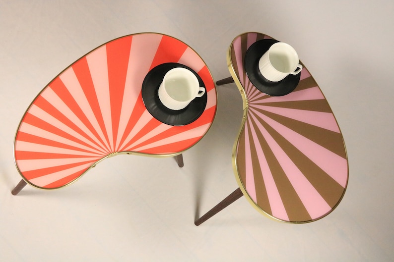 May include: Two kidney-shaped side tables with dark wooden legs and gold-toned metal rims. One table features a red and pink sunburst pattern, while the other has a brown and pink sunburst pattern. Each table holds a black saucer and white cup.