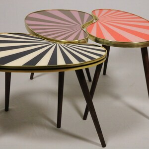 May include: Set of three mid-century modern accent tables. Each table has a unique oval shape with a sunburst design in black and white, pink and gray, and coral and pink. The tables have brass trim and dark brown wooden legs.