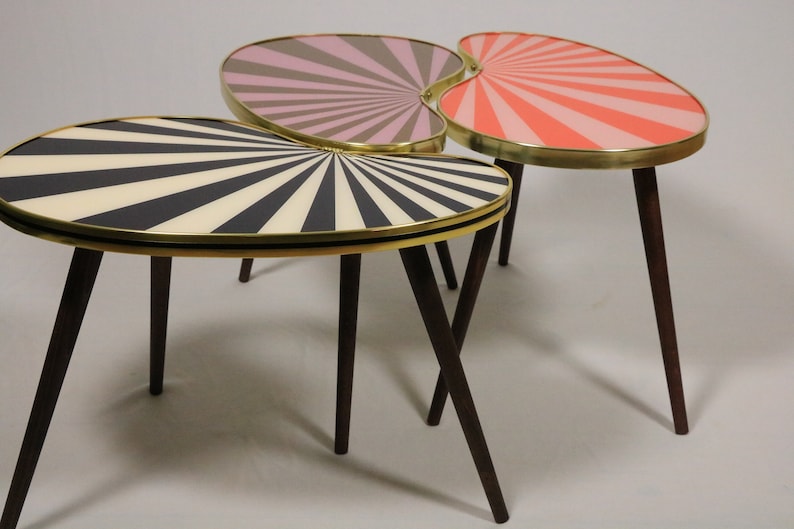 May include: Set of three mid-century modern accent tables. Each table has a unique oval shape with a sunburst design in black and white, pink and gray, and coral and pink. The tables have brass trim and dark brown wooden legs.