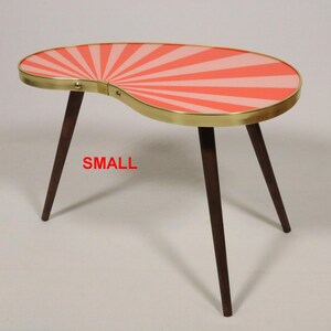 May include: A kidney-shaped side table with three dark brown tapered wooden legs and a gold-toned metal rim. The tabletop features a sunburst pattern in alternating shades of coral and light pink. The word SMALL is printed in red.