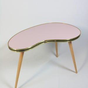 Puede incluir: Una mesa de centro en forma de riñón de estilo moderno de mediados de siglo. La mesa tiene una tapa rosa claro con un borde dorado y un acento negro. Tiene tres patas de madera cónicas. Mide unos 90 cm de largo.