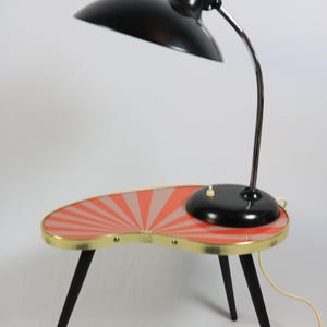 May include: A mid-century modern accent table with a built-in lamp. The table has a kidney-shaped top with a red and pink sunburst design, brass trim, and black legs. The lamp has a black shade and a chrome gooseneck.